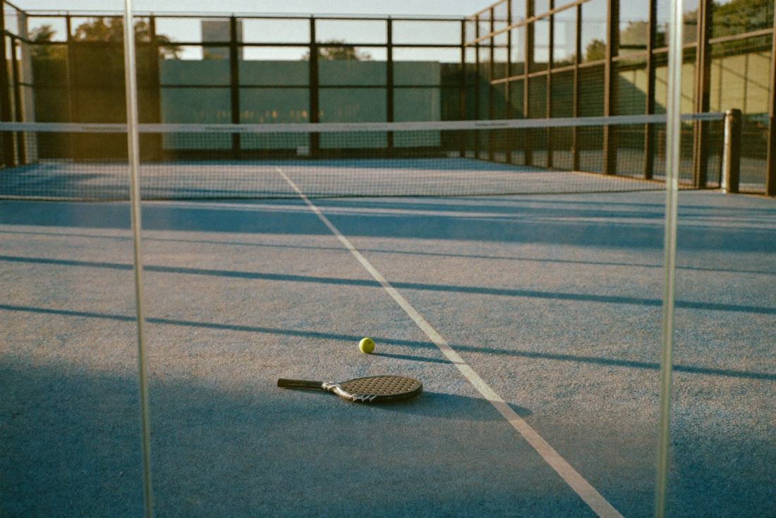 Een leeg padelterrein met blauw kunstgras en glazen wanden, een padelracket en een bal op de grond.